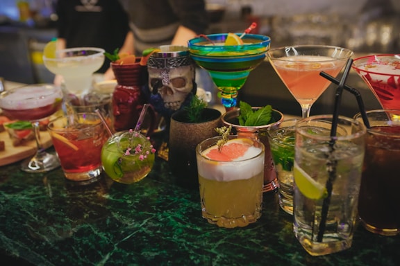 A variety of colorful cocktails arranged on a bar counter, each in a distinct glass type. There are garnishes like fruit slices, mint leaves, and decorative flowers. The setting appears to be a lively bar environment with a mix of classic and tiki-themed drinkware, including a skull-shaped mug.