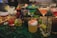 A variety of colorful cocktails arranged on a bar counter, each in a distinct glass type. There are garnishes like fruit slices, mint leaves, and decorative flowers. The setting appears to be a lively bar environment with a mix of classic and tiki-themed drinkware, including a skull-shaped mug.
