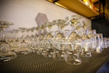 Many clean and clear wine glasses and other types of glasses are seen arranged upside down on a black textured mat. The background shows a shelf with various items and a wall with a geometric pattern.