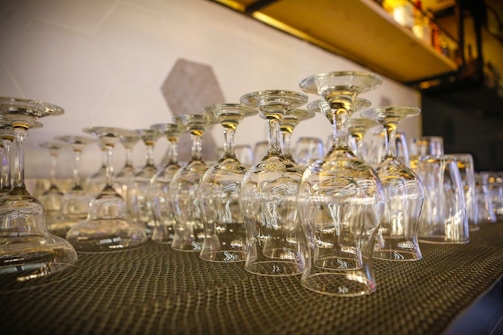 Many clean and clear wine glasses and other types of glasses are seen arranged upside down on a black textured mat. The background shows a shelf with various items and a wall with a geometric pattern.