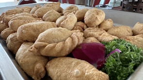 A tray filled with golden-brown fried pastries arranged closely together. They have a crispy, flaky appearance with twisted edges, suggesting they might be empanadas or similar baked goods. Accompanying the pastries are garnishes of bright purple petals and fresh green parsley.