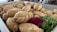 A tray filled with golden-brown fried pastries arranged closely together. They have a crispy, flaky appearance with twisted edges, suggesting they might be empanadas or similar baked goods. Accompanying the pastries are garnishes of bright purple petals and fresh green parsley.