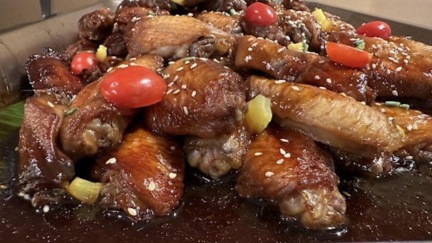 A mouth-watering platter of glazed chicken wings adorned with juicy cherry tomatoes and sprinkled with sesame seeds. The wings are garnished with chunks of pineapple and fresh herbs on a dark sauce base.