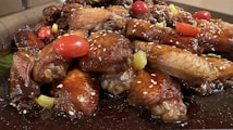 A mouth-watering platter of glazed chicken wings adorned with juicy cherry tomatoes and sprinkled with sesame seeds. The wings are garnished with chunks of pineapple and fresh herbs on a dark sauce base.