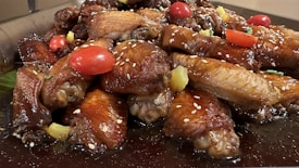 A mouth-watering platter of glazed chicken wings adorned with juicy cherry tomatoes and sprinkled with sesame seeds. The wings are garnished with chunks of pineapple and fresh herbs on a dark sauce base.