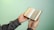 A person is holding an open book with Arabic script, possibly a religious text, against a smooth, solid green background. The person's sleeves are checkered, and their hands are gently gripping the book.