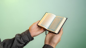 A person is holding an open book with Arabic script, possibly a religious text, against a smooth, solid green background. The person's sleeves are checkered, and their hands are gently gripping the book.