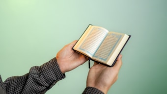 A person is holding an open book with Arabic script, possibly a religious text, against a smooth, solid green background. The person's sleeves are checkered, and their hands are gently gripping the book.