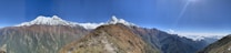 A panoramic view of a mountain range featuring snow-capped peaks and rugged terrain. The foreground shows a rocky, grassy path leading up the slope while the background reveals expansive valleys and ridges of varying elevations. The sky is clear and bright with the sun shining intensely from the top right corner of the image. Below the distant peaks, there are layers of clouds floating just above the horizon.