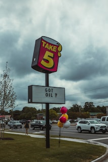A road sign advertising 'Take 5' with the phrase 'GOT OIL ?' displayed beneath it. The sign is positioned by a roadside with several vehicles on the street, and a person walking on the sidewalk. Balloons in pink, yellow, and white are attached to the signpost. The sky is overcast, with gray clouds in the background.