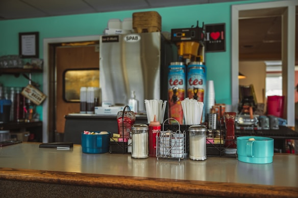 Countertop with various condiments and utensils including straws, napkins, a ketchup bottle, sugar packets, and a teal cup. Behind, there are drink dispensers labeled 'tea' and other kitchen items. The walls are painted a light blue, and there is a framed sign and a decorative shelf.