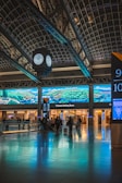 A train station interior featuring a large digital screen displaying landscape graphics. The space includes a clock suspended from the ceiling, a reflective floor, and several passengers with luggage facing the ticketed waiting room area.