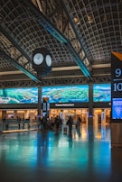 A train station interior featuring a large digital screen displaying landscape graphics. The space includes a clock suspended from the ceiling, a reflective floor, and several passengers with luggage facing the ticketed waiting room area.