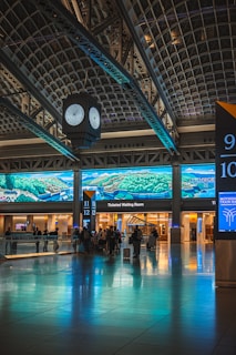 A train station interior featuring a large digital screen displaying landscape graphics. The space includes a clock suspended from the ceiling, a reflective floor, and several passengers with luggage facing the ticketed waiting room area.