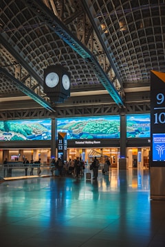 A train station interior featuring a large digital screen displaying landscape graphics. The space includes a clock suspended from the ceiling, a reflective floor, and several passengers with luggage facing the ticketed waiting room area.