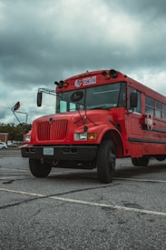A large red bus is parked in an empty parking lot under a cloudy sky. The bus has 'Martial Arts World' written on the front and the license plate shows a registration from Virginia. The vehicle is positioned on a cracked asphalt surface, and the surrounding area appears quiet.