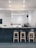A minimalist cafe interior with a sleek, modern bar counter in a muted dark color. Four wooden stools are neatly aligned in front of the counter. The background features a coffee machine and various jars, suggesting a coffee shop environment. Overhead, a contemporary light fixture provides illumination.