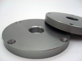 Two round metallic plates, each with holes and a central threaded hole, laying on a flat surface. The plates have a smooth, polished finish with a grayish metallic color.