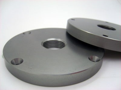 Two round metallic plates, each with holes and a central threaded hole, laying on a flat surface. The plates have a smooth, polished finish with a grayish metallic color.