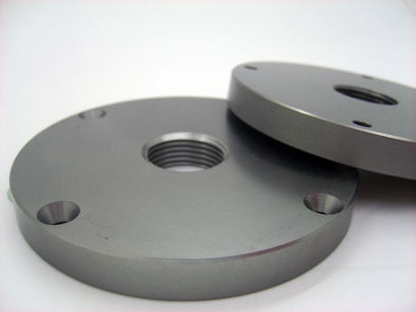 Two round metallic plates, each with holes and a central threaded hole, laying on a flat surface. The plates have a smooth, polished finish with a grayish metallic color.