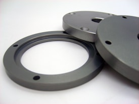 Two metallic circular components with multiple holes, possibly industrial parts, laid out on a flat, light-colored surface. They have a smooth, polished appearance, indicating precision manufacturing.