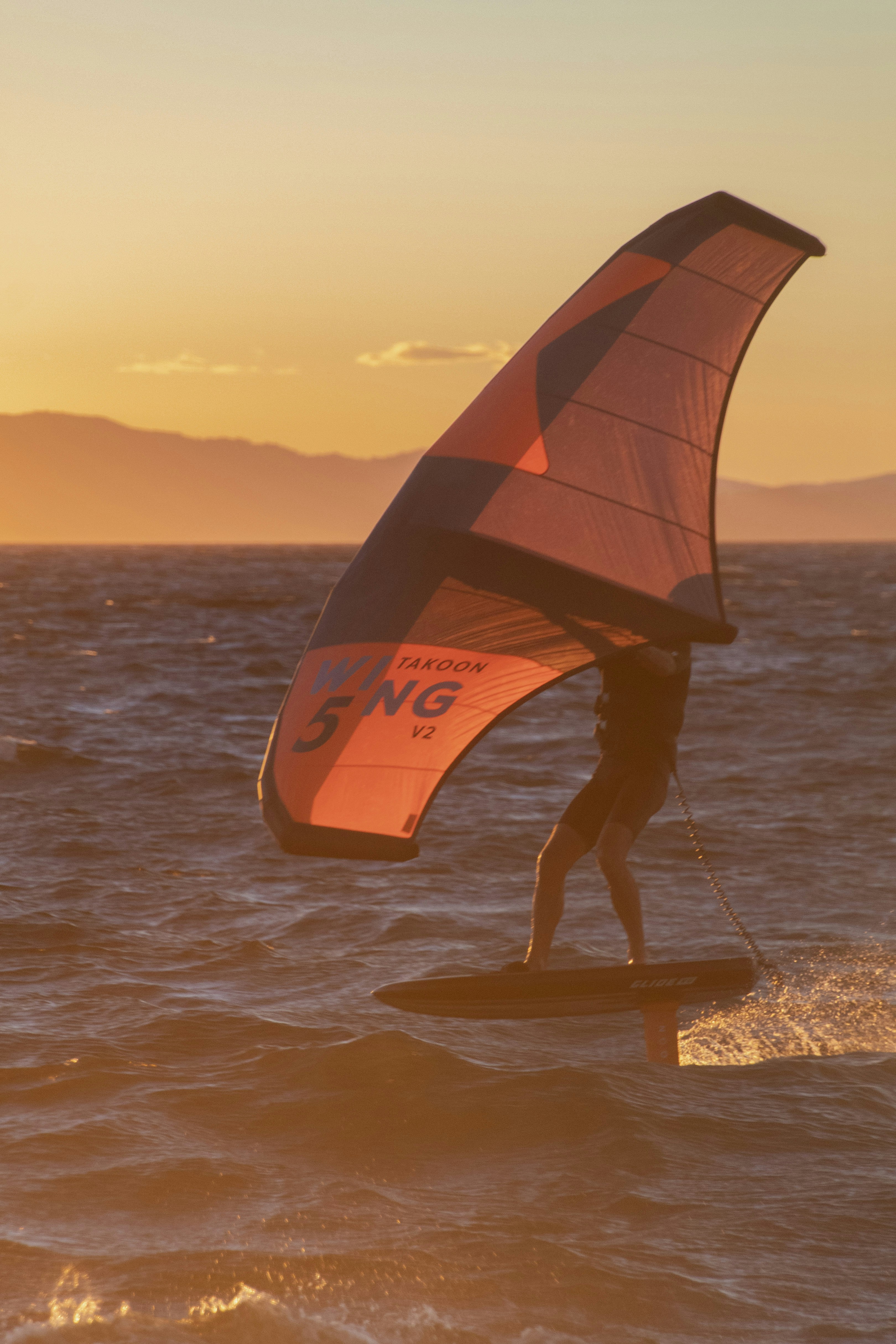 Persona practicando wing foil en el agua al atardecer con montañas.