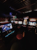A dimly lit arcade room with several retro arcade game machines lined up. The machines display various game screens, and there are round stools in front of them. The atmosphere is nostalgic, with the soft glow of the screens providing the primary illumination.