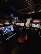 A dimly lit arcade room with several retro arcade game machines lined up. The machines display various game screens, and there are round stools in front of them. The atmosphere is nostalgic, with the soft glow of the screens providing the primary illumination.