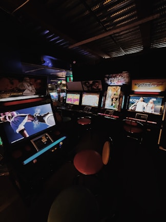 A dimly lit arcade room with several retro arcade game machines lined up. The machines display various game screens, and there are round stools in front of them. The atmosphere is nostalgic, with the soft glow of the screens providing the primary illumination.