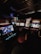 A dimly lit arcade room with several retro arcade game machines lined up. The machines display various game screens, and there are round stools in front of them. The atmosphere is nostalgic, with the soft glow of the screens providing the primary illumination.