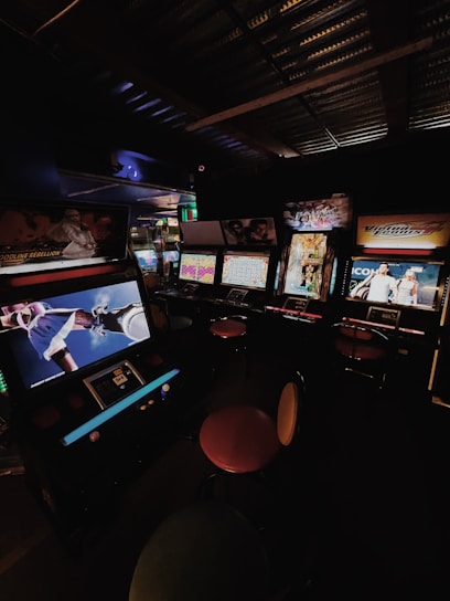 A dimly lit arcade room with several retro arcade game machines lined up. The machines display various game screens, and there are round stools in front of them. The atmosphere is nostalgic, with the soft glow of the screens providing the primary illumination.