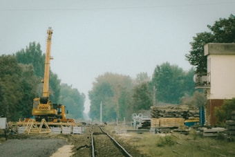 A railway construction site with tracks extending into the distance. There is a yellow crane with marked lettering situated on the tracks. Piles of building materials and wooden pallets are stacked nearby. The area is surrounded by dense trees and there is a small building on the right side.