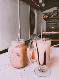 Two beverages in glass mugs with metal straws rest on a table. The drink on the left is a creamy pinkish color with ice, served in a mason jar. The drink on the right has a similar color but is topped with dark chocolate drizzle. In the background, the interior of a rustic café is visible, featuring wooden shelves and a menu board.