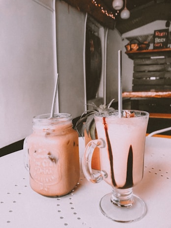 Two beverages in glass mugs with metal straws rest on a table. The drink on the left is a creamy pinkish color with ice, served in a mason jar. The drink on the right has a similar color but is topped with dark chocolate drizzle. In the background, the interior of a rustic café is visible, featuring wooden shelves and a menu board.
