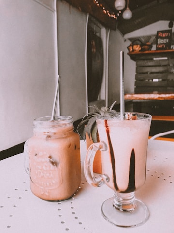 Two beverages in glass mugs with metal straws rest on a table. The drink on the left is a creamy pinkish color with ice, served in a mason jar. The drink on the right has a similar color but is topped with dark chocolate drizzle. In the background, the interior of a rustic café is visible, featuring wooden shelves and a menu board.