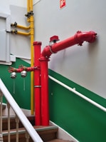 A bright red firefighting pipe system is installed along a staircase with green and white walls. The pipes include prominent valves and connectors, and a yellow gas pipe runs parallel to the firefighting system. The stairs have wooden steps and white railings.