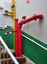 A bright red firefighting pipe system is installed along a staircase with green and white walls. The pipes include prominent valves and connectors, and a yellow gas pipe runs parallel to the firefighting system. The stairs have wooden steps and white railings.