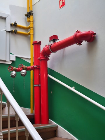 A bright red firefighting pipe system is installed along a staircase with green and white walls. The pipes include prominent valves and connectors, and a yellow gas pipe runs parallel to the firefighting system. The stairs have wooden steps and white railings.