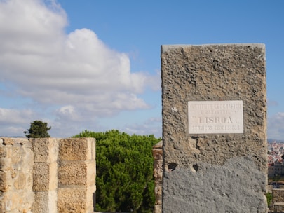 A stone marker with an inscription in Portuguese stands prominently against a backdrop of trees and a clear blue sky with scattered white clouds. The plaque on the marker mentions the Instituto Geográfico e Cadastral in Lisbon. In the distance, urban structures peek through the foliage.