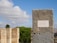 A stone marker with an inscription in Portuguese stands prominently against a backdrop of trees and a clear blue sky with scattered white clouds. The plaque on the marker mentions the Instituto Geográfico e Cadastral in Lisbon. In the distance, urban structures peek through the foliage.