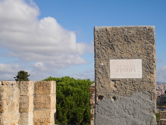 A stone marker with an inscription in Portuguese stands prominently against a backdrop of trees and a clear blue sky with scattered white clouds. The plaque on the marker mentions the Instituto Geogr&aacute;fico e Cadastral in Lisbon. In the distance, urban structures peek through the foliage.