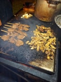 Grilled meat skewers and sliced mushrooms are cooking on a flat metal grill. A large cooking pot is partially visible in the background, and a plate is on the side. The setting appears to be outdoors, with a warm, inviting atmosphere.