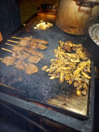 Grilled meat skewers and sliced mushrooms are cooking on a flat metal grill. A large cooking pot is partially visible in the background, and a plate is on the side. The setting appears to be outdoors, with a warm, inviting atmosphere.