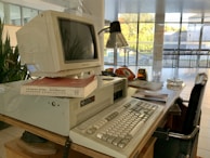 A retro computer setup is on a wooden desk, featuring a bulky Packard Bell monitor sitting on an elevated base. Nearby are books, including one with a red cover. The desk also includes an old-fashioned orange rotary phone, a lamp, papers, a glass ashtray, and an analogue clock with an orange casing. The scene is set in an environment with large glass windows, allowing natural light to brighten the area.