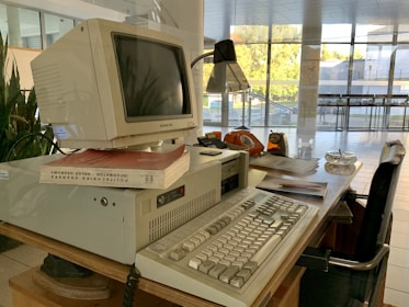 A retro computer setup is on a wooden desk, featuring a bulky Packard Bell monitor sitting on an elevated base. Nearby are books, including one with a red cover. The desk also includes an old-fashioned orange rotary phone, a lamp, papers, a glass ashtray, and an analogue clock with an orange casing. The scene is set in an environment with large glass windows, allowing natural light to brighten the area.