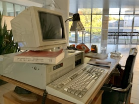 A retro computer setup is on a wooden desk, featuring a bulky Packard Bell monitor sitting on an elevated base. Nearby are books, including one with a red cover. The desk also includes an old-fashioned orange rotary phone, a lamp, papers, a glass ashtray, and an analogue clock with an orange casing. The scene is set in an environment with large glass windows, allowing natural light to brighten the area.