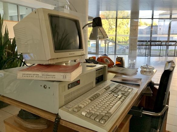 A retro computer setup is on a wooden desk, featuring a bulky Packard Bell monitor sitting on an elevated base. Nearby are books, including one with a red cover. The desk also includes an old-fashioned orange rotary phone, a lamp, papers, a glass ashtray, and an analogue clock with an orange casing. The scene is set in an environment with large glass windows, allowing natural light to brighten the area.