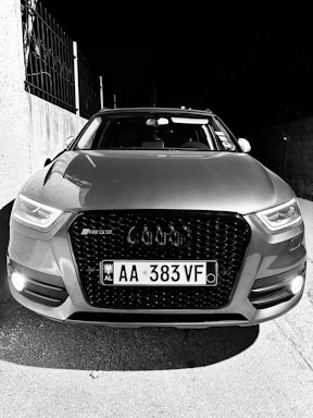 The image features the front view of a car with a prominent grille and headlights. The vehicle displays a license plate with the code 'AA 383 VF'. The surrounding area includes a concrete wall and a metal fence on the left side.