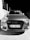 The image features the front view of a car with a prominent grille and headlights. The vehicle displays a license plate with the code 'AA 383 VF'. The surrounding area includes a concrete wall and a metal fence on the left side.