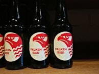 Several dark-colored beer bottles are displayed with labels featuring a red and white stylized eagle head and the text 'FALKEN BIER' along with 'LAGER HELL' printed on them. The bottles are neatly arranged in a row on a wooden shelf.